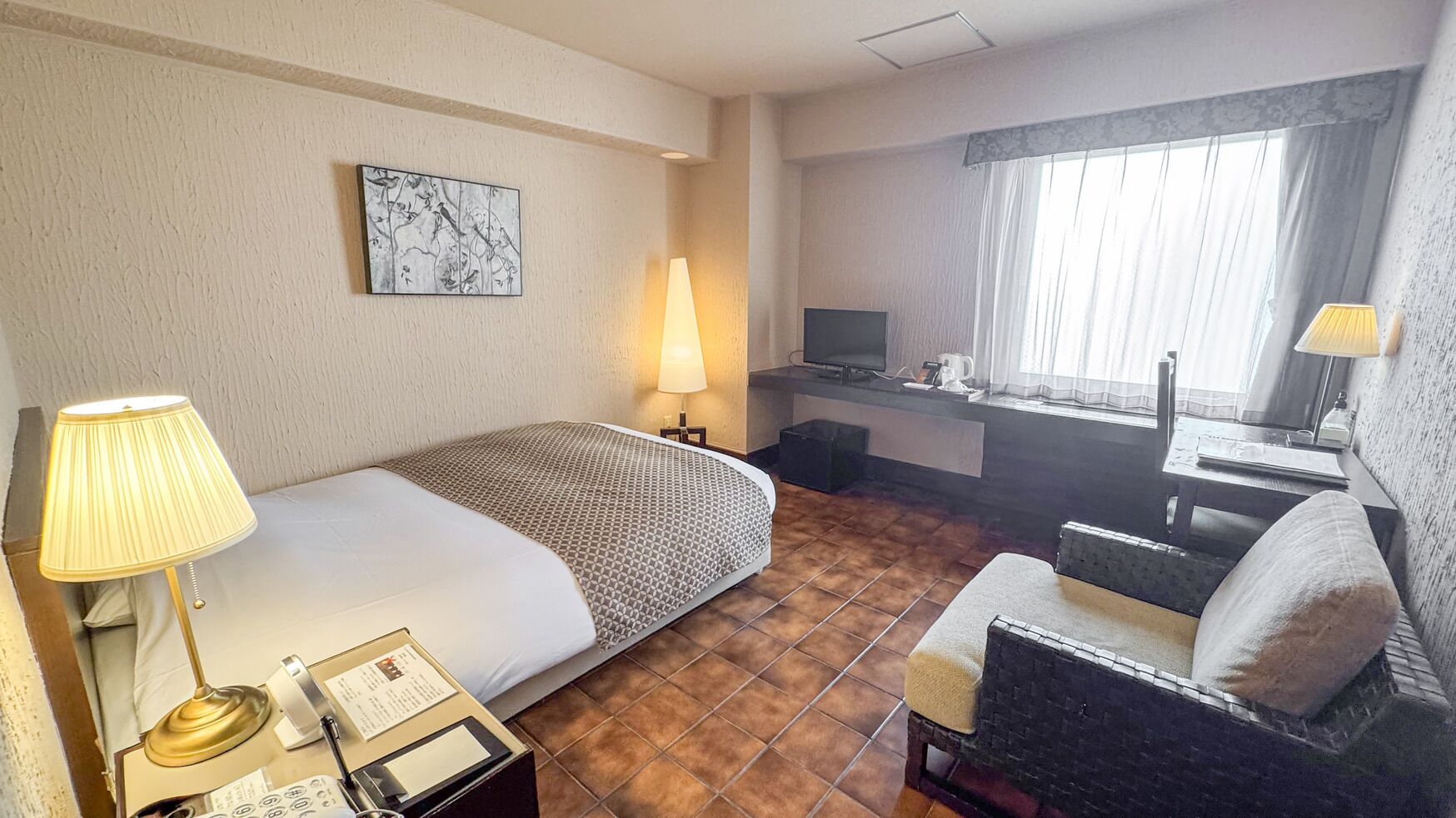 Ein Hotelzimmer mit Bett, Schreibtisch, Stuhl, Lampe, Fernseher und einem Fenster mit Vorhängen.