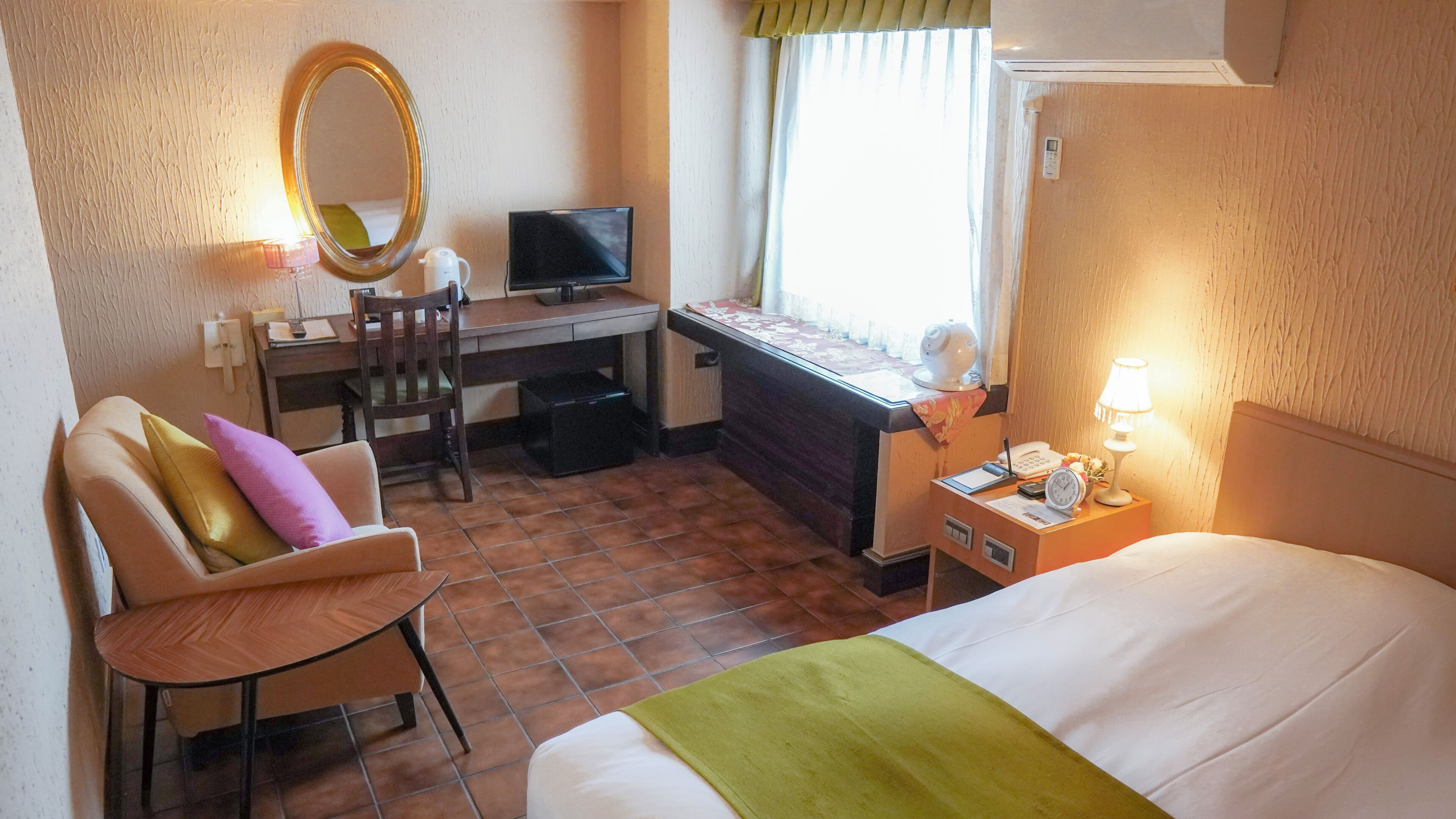 A hotel room with a bed, a desk with a computer, a chair, a mirror, a bedside table with a lamp, and a window with curtains.