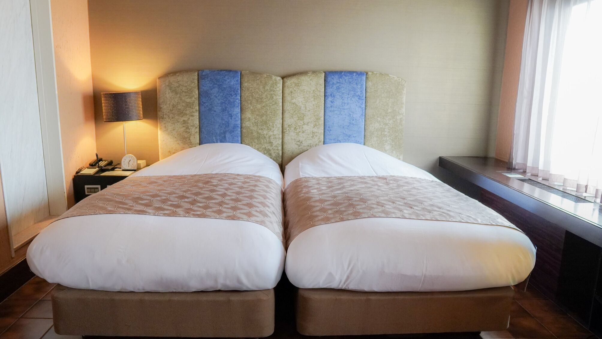 Ein Doppelbett mit gemustertem Überwurf, ein Nachttisch mit Lampe und Blick ins Freie durch ein Fenster.