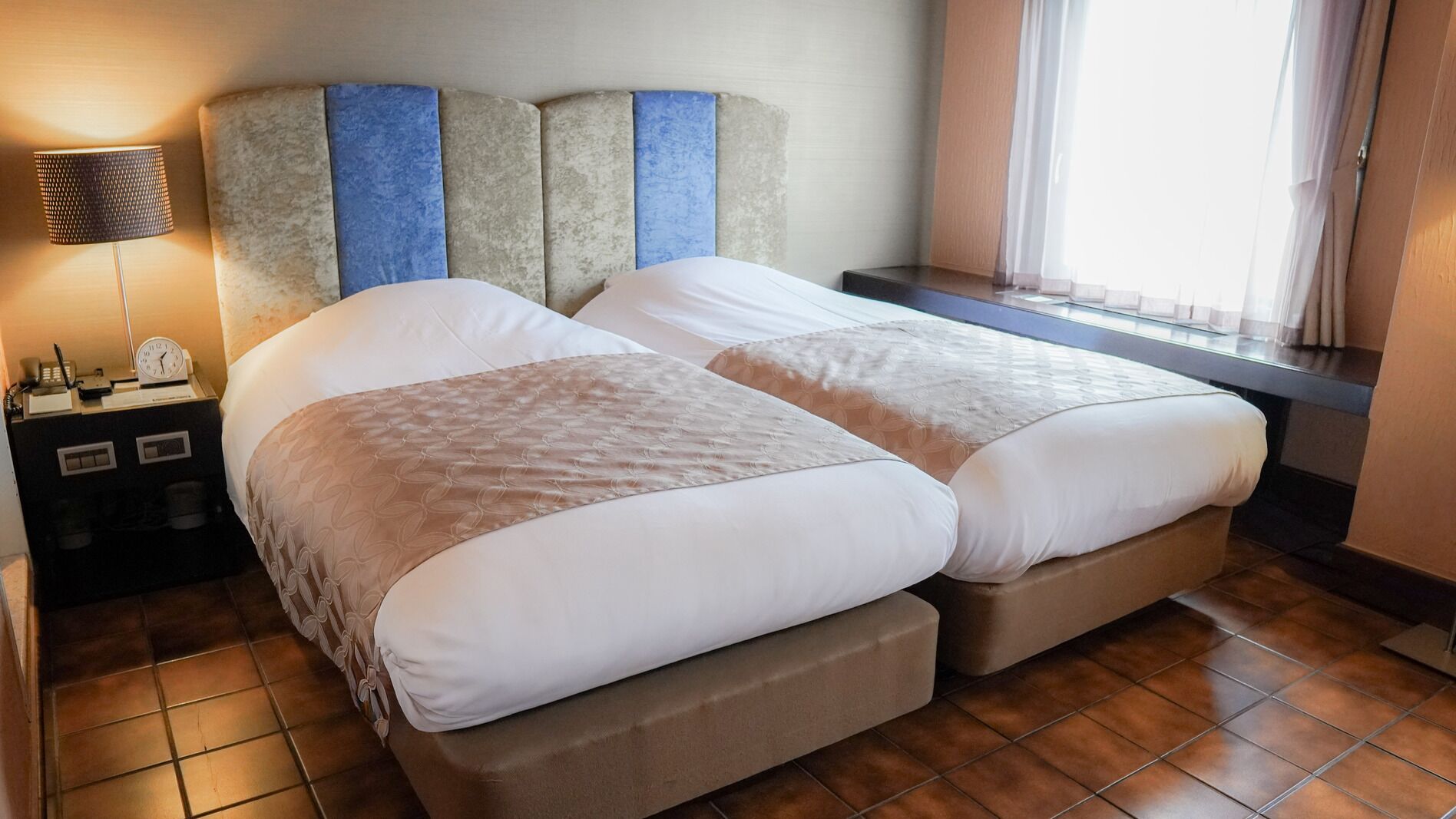 Ein Doppelbett mit Kopfteil, ein Nachttisch mit Lampe und ein Fenster mit Vorhängen.