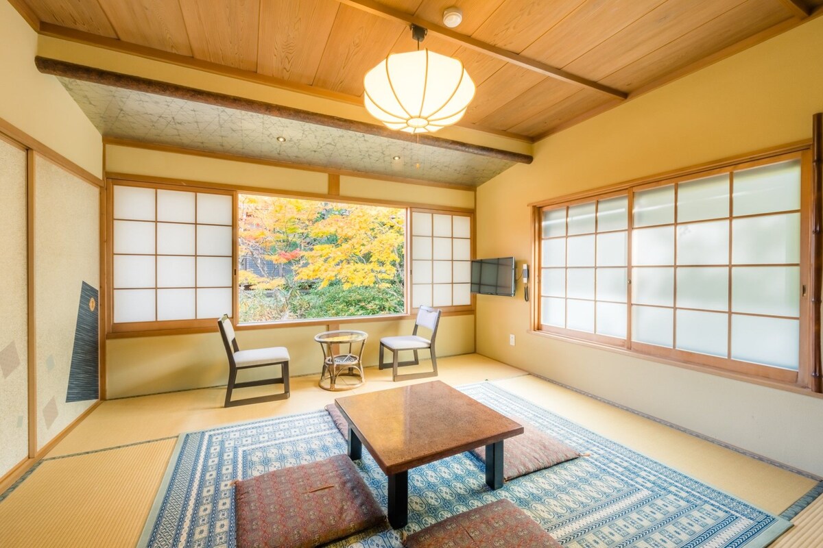 A traditional Japanese room with tatami flooring, a low wooden table, two chairs, a shoji screen, and a view of autumn foliage through a large window.