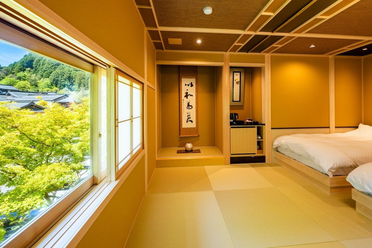 Quarto japonês tradicional com tatames, uma cama, uma pequena mesa e vista para uma área arborizada.
