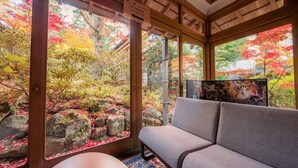 A room with a view of a garden with autumn foliage, a sofa, and a television. - Ekoin (Koya)