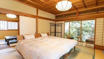 Un dormitorio de estilo japonés tradicional con suelo de tatami, una cama grande, un escritorio y vistas a la vegetación a través de puertas correderas.