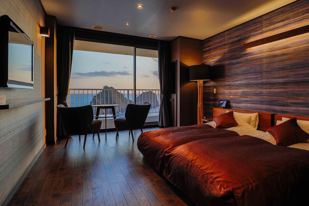 A modern hotel room with a large bed, a balcony with a table and chairs, and a view of the sea.