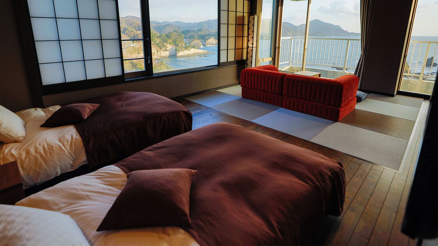 A room with two beds, a red sofa, tatami mats, and a large window with a view of the sea and mountains.