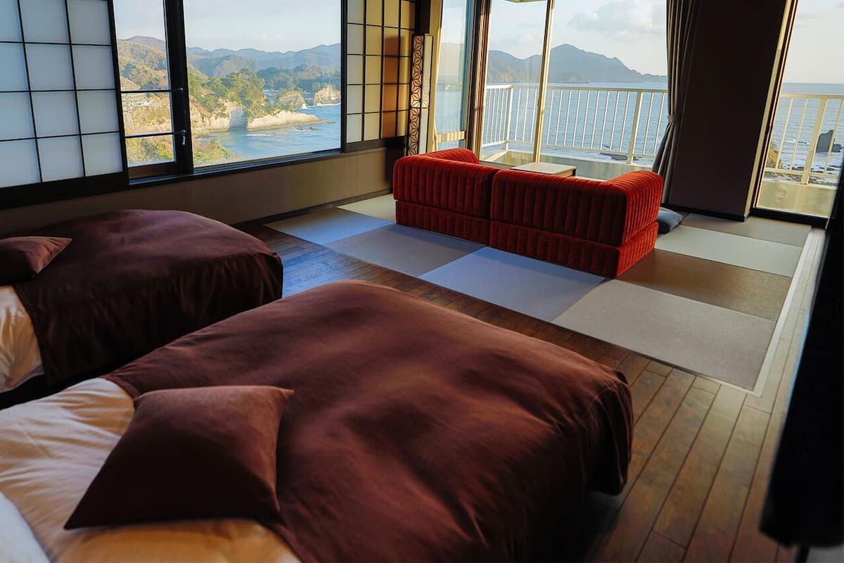 A room with two beds, a red sofa, tatami mats, and a large window with a view of the sea and mountains.