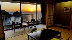 A traditional Japanese room with tatami flooring, a low table, and chairs. A large window offers a view of the sea and sunset. - Dogashima Hotel Tenyu (Nishiizu)