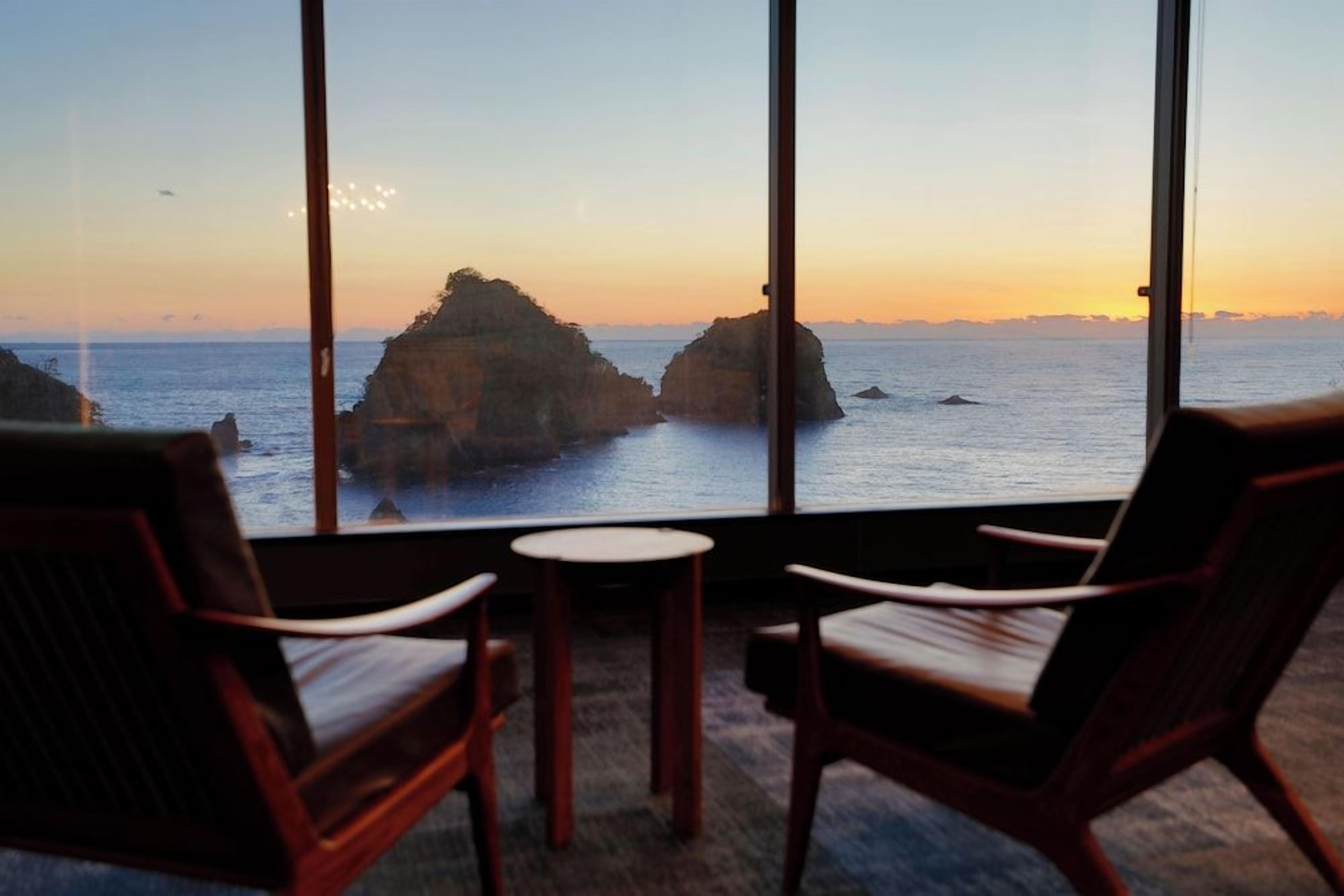 A room with a large window overlooking a coastal landscape with rock formations and the sea. There are two chairs and a small table in the room.