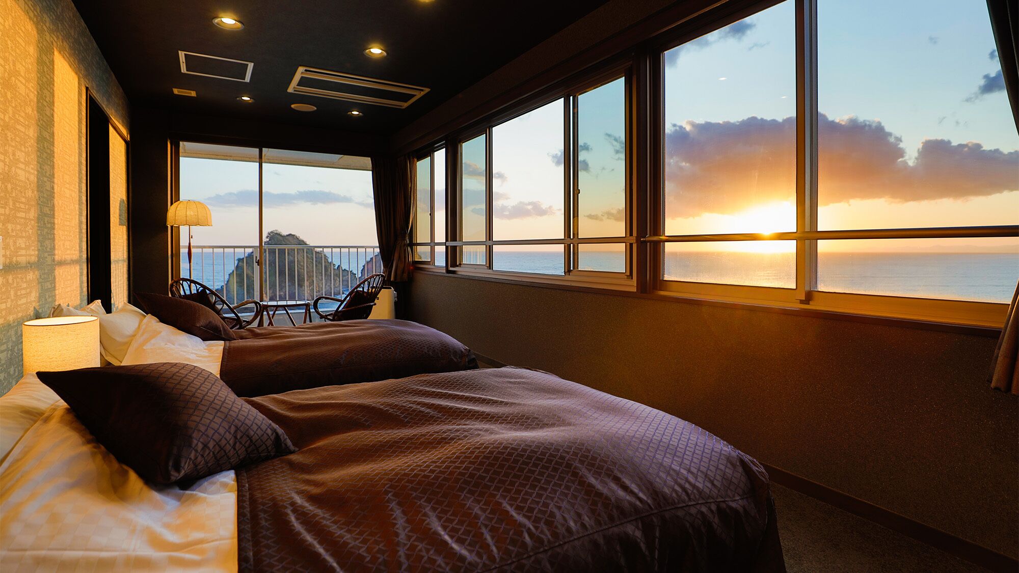 A room with a bed, a chair, a lamp, and a large window with a sunset view.