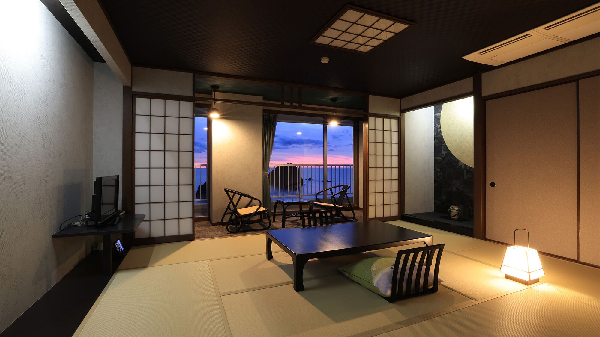 Een traditionele Japanse kamer met tatami vloerbedekking, een lage tafel, stoelen, een televisie en uitzicht op de zonsondergang door een groot raam.