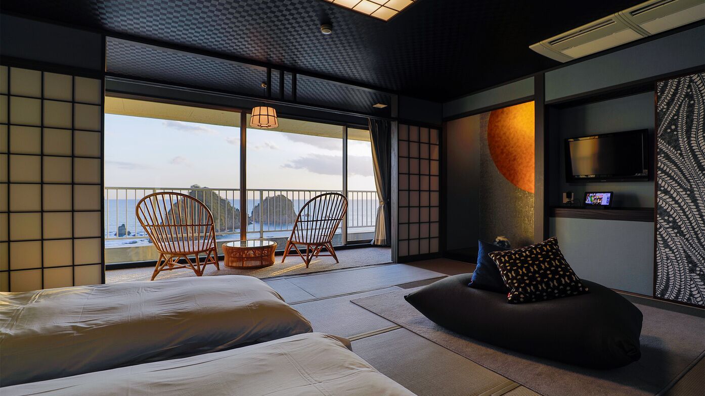 A modern hotel room with a large window, a bed, a seating area, and a TV.