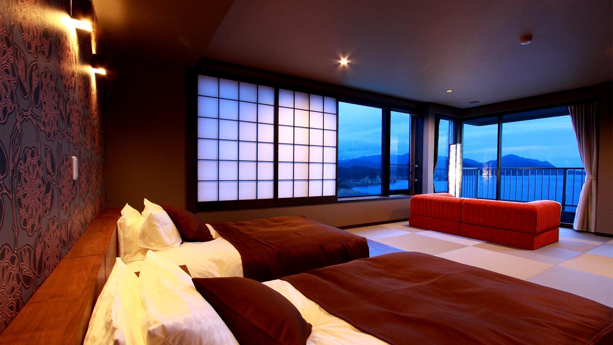 Een hotelkamer met twee bedden, een rode bank, grote ramen en uitzicht, en een kamer met een patroon op de muur.