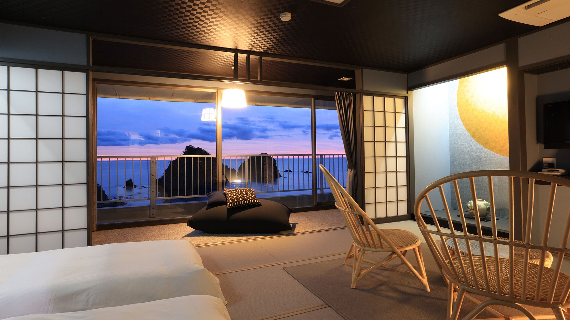 A room with a bed, a chair, a table, and a view of the sea at dusk.