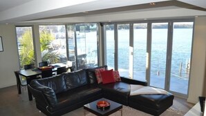 A modern living room with a black leather sofa, a coffee table, and large windows overlooking a body of water. - Bancroft Views Apartments (Metung)