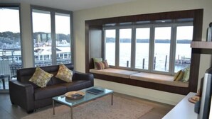 A living room with a sofa, a coffee table, and a window seat overlooking a body of water. - Bancroft Views Apartments (Metung)
