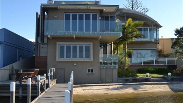 A modern two-story building with a balcony, a dock, and a landscaped area with a palm tree. - Bancroft Views Apartments (Metung)