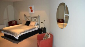 A bedroom with a bed, a chair, a small table, a nightstand, a lamp, a mirror, and a wall art piece. - Bancroft Views Apartments (Metung)
