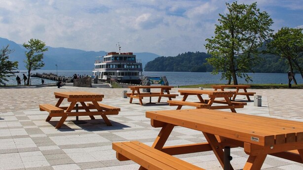 A lakeside picnic area with wooden tables and benches, a dock, and a boat in the background. - Towadako Lakeview Hotel (Kosaka)