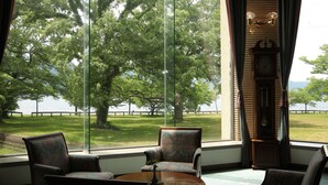 A room with a large window overlooking a grassy area and a body of water. There is a wooden clock on the wall and a chair with a vase on a table in the foreground. - Towadako Lakeview Hotel (Kosaka)