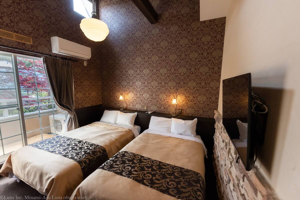 Habitación de hotel con dos camas, un ventanal con cortinas, un equipo de aire acondicionado y un televisor instalado en la pared.