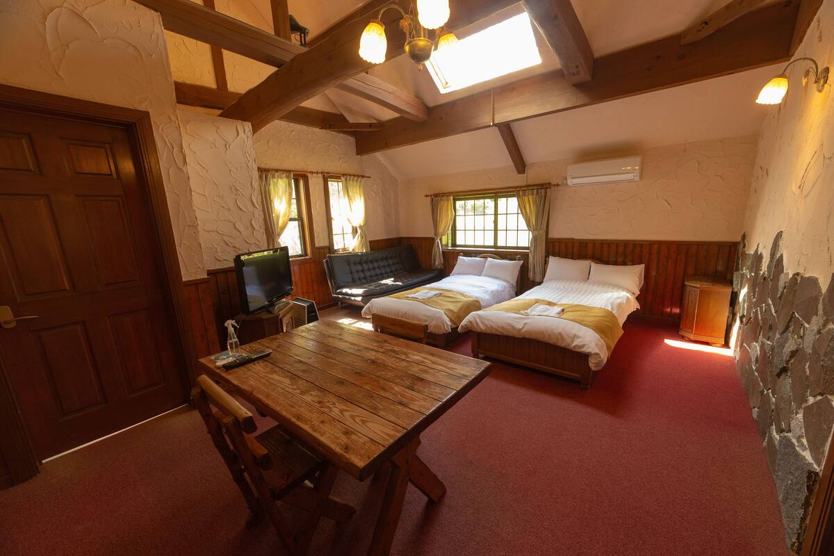 Una habitación con una mesa de madera, dos camas, un televisor y una pared de piedra.
