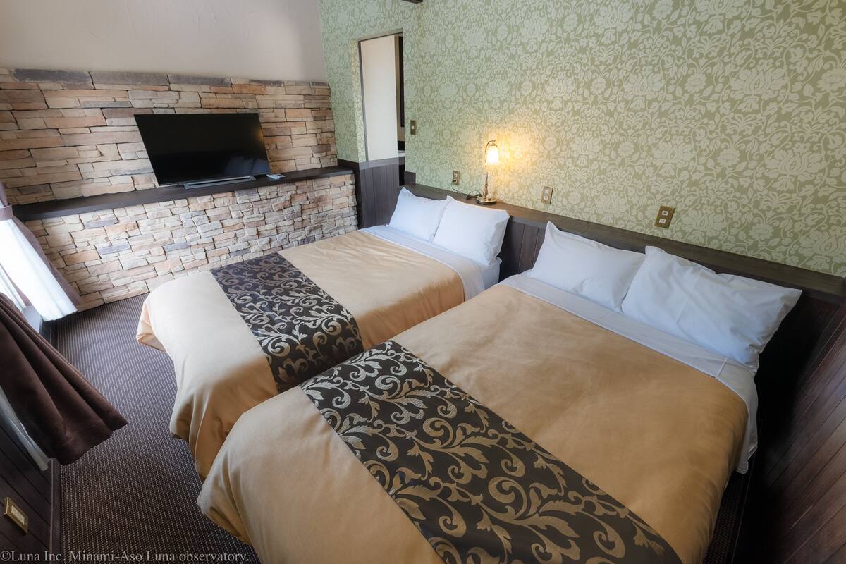 Habitación de hotel con dos camas, un televisor montado en una pared de ladrillo y un papel tapiz estampado.