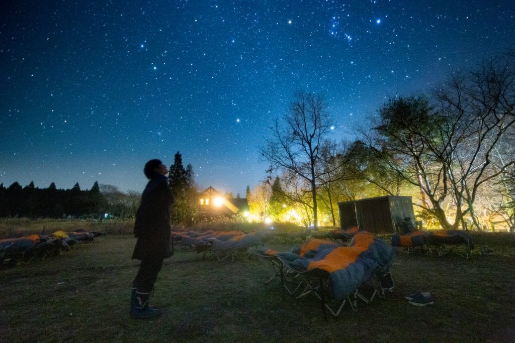 Una persona bajo un cielo estrellado, con equipo de campamento y una casa al fondo.