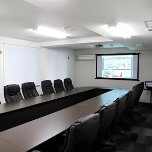 A conference room with a long table and six chairs, a projector screen, and an air conditioning unit on the wall.