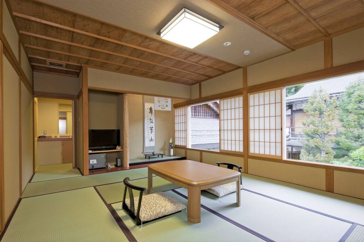 A traditional Japanese room with tatami flooring, a low table, a television, and a view of a residential area.