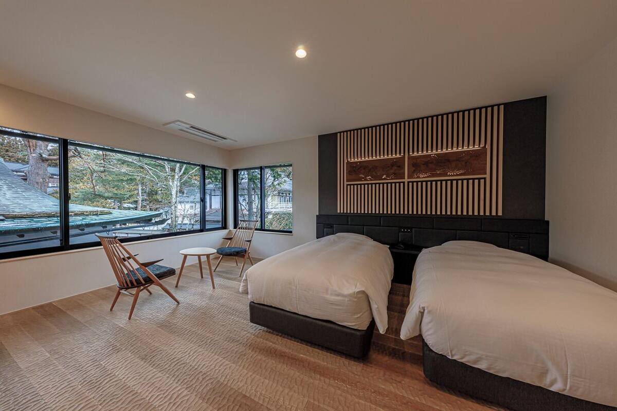 A modern hotel room with two beds, a large window, a wooden chair, and a small table.