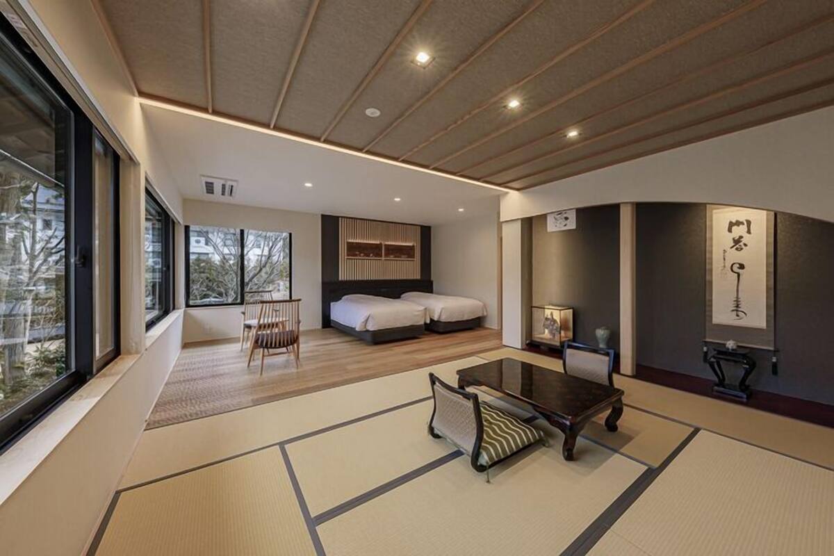 A modern hotel room with a large window, a bed, a dining table, and a traditional Japanese tatami mat floor.