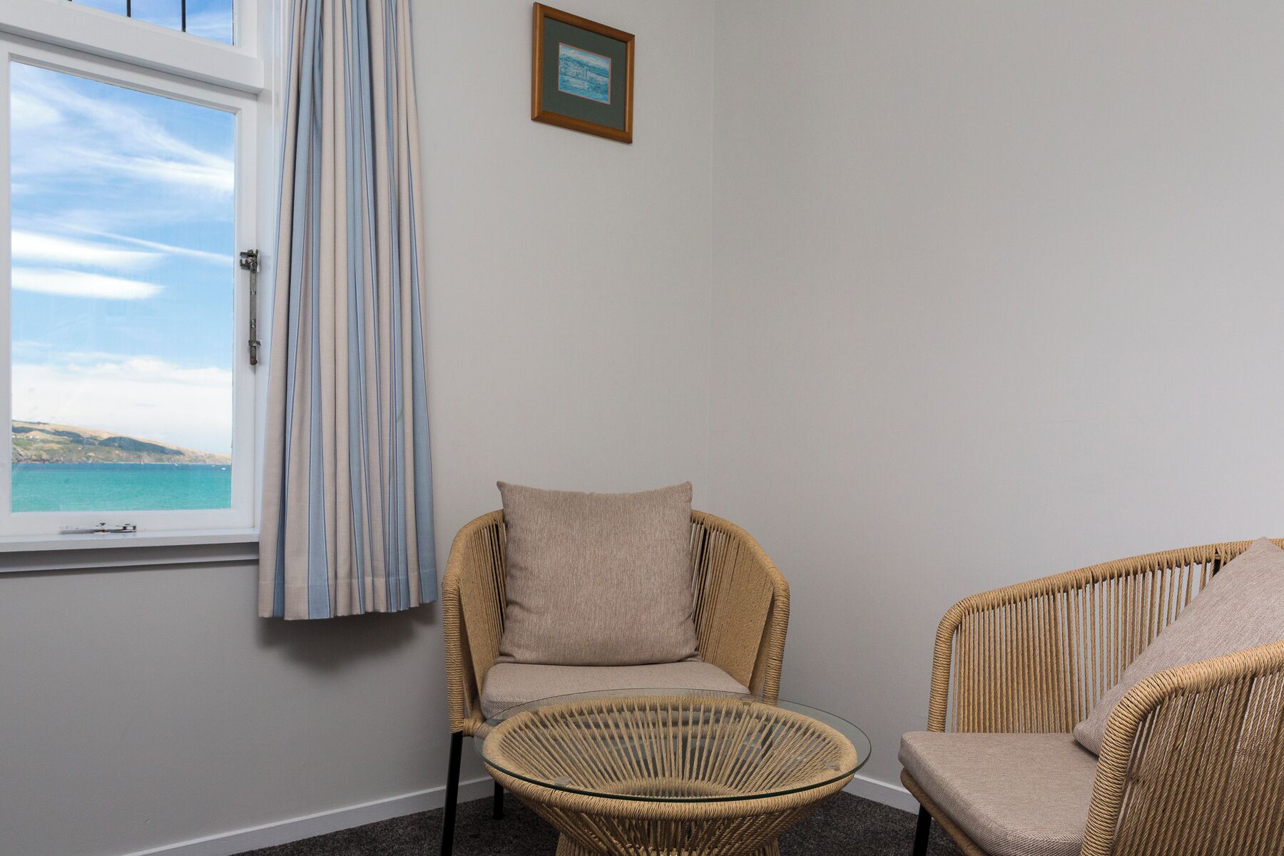 A room with a wicker chair and a small glass table, a window with a view of the sea, and a framed picture on the wall.