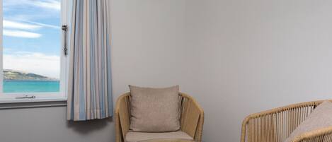A room with a wicker chair and a small glass table, a window with a view of the sea, and a framed picture on the wall.
