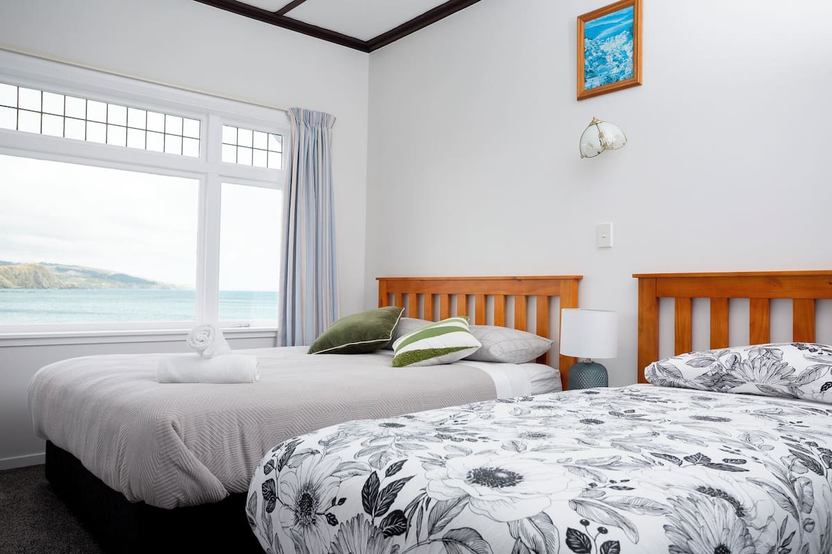 A hotel room with two beds, a large window with a view of the sea, a framed picture on the wall, and a small table with a lamp.