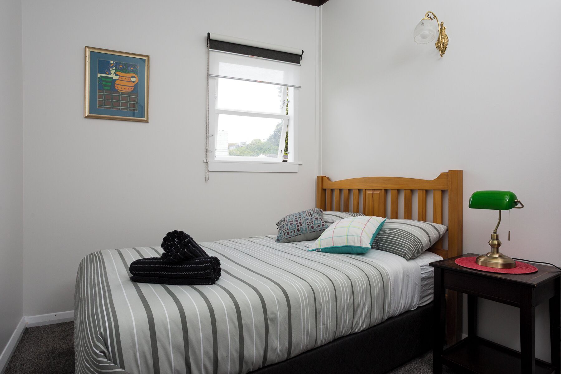 A bedroom with a bed, a nightstand, a lamp, a window with blinds, and a framed picture on the wall.