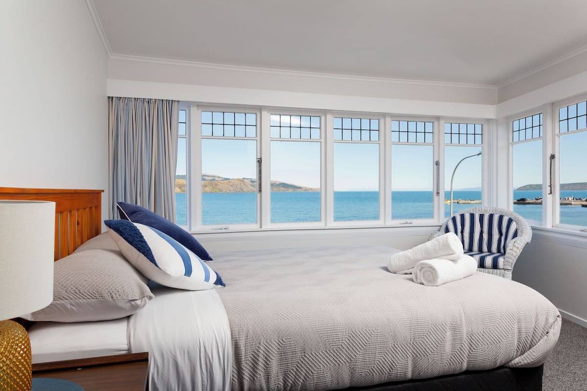 A bedroom with a large window overlooking a coastal view, a bed with a striped chair, and a wooden headboard.
