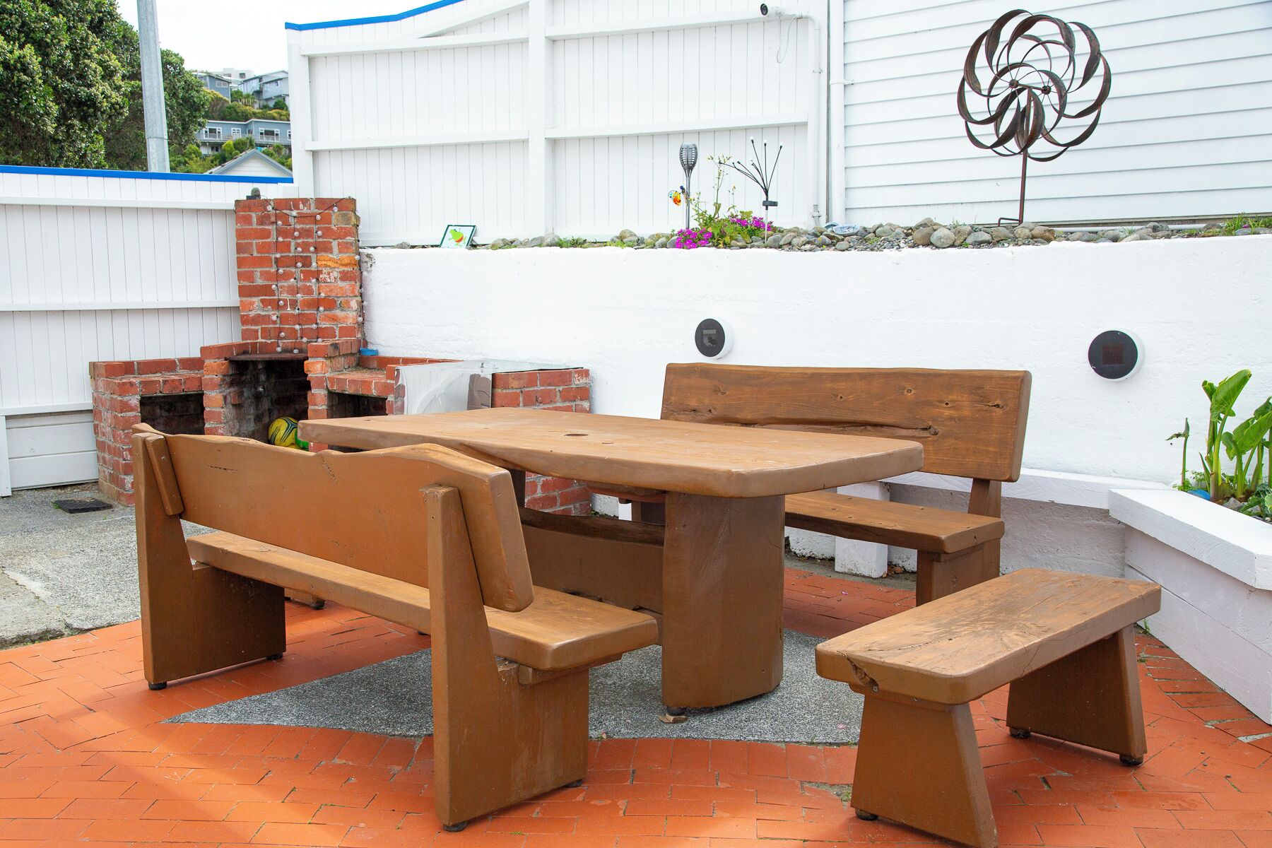 Um pátio externo com mesa e bancos de madeira, lareira de tijolos e uma escultura metálica decorativa.