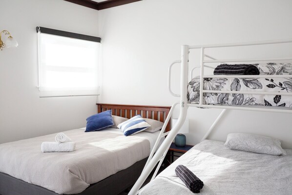 Habitación con dos camas, una literas y otra individual. Cuenta con ventana con persiana, mesita de noche con lámpara, y un aplique en la pared.