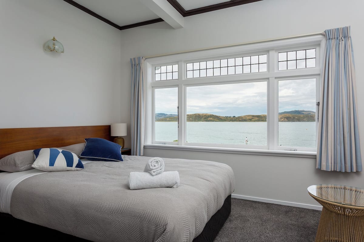 A bedroom with a large window, a bed with a headboard, a bedside table, and a lamp.