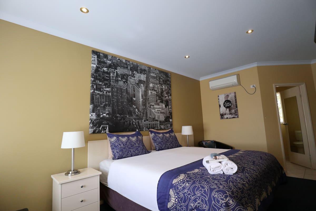 A hotel room with a bed, two bedside tables, a wall-mounted air conditioner, and a door leading to the bathroom.
