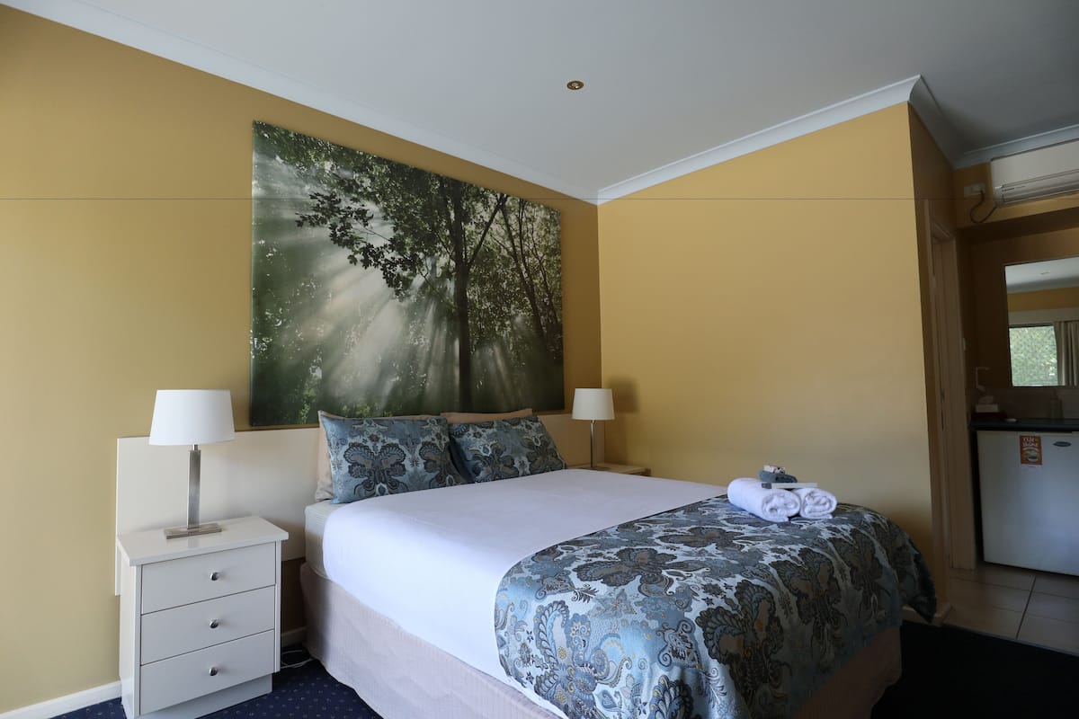 A hotel room with a bed, two bedside lamps, a nightstand, a large wall painting, and a refrigerator.