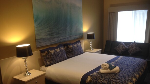Une chambre d’hôtel avec un grand lit, deux tables de chevet avec des lampes, un tableau représentant une vague accroché au mur et une fenêtre avec des rideaux.