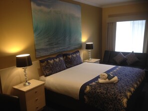 A hotel room with a large bed, two bedside tables with lamps, a painting of a wave on the wall, and a window with curtains. - Kyabram Motor Inn (Kyabram)