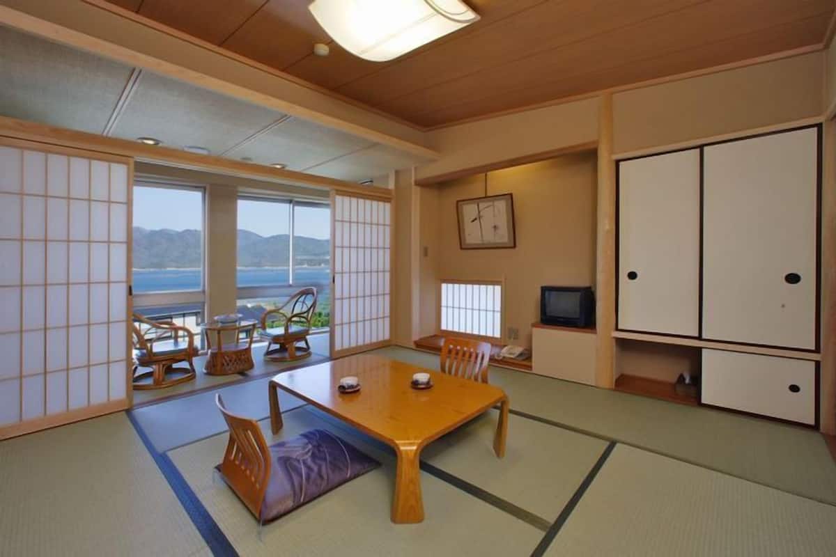 A traditional Japanese room with tatami flooring, a wooden table, chairs, a TV, and a view of the sea.
