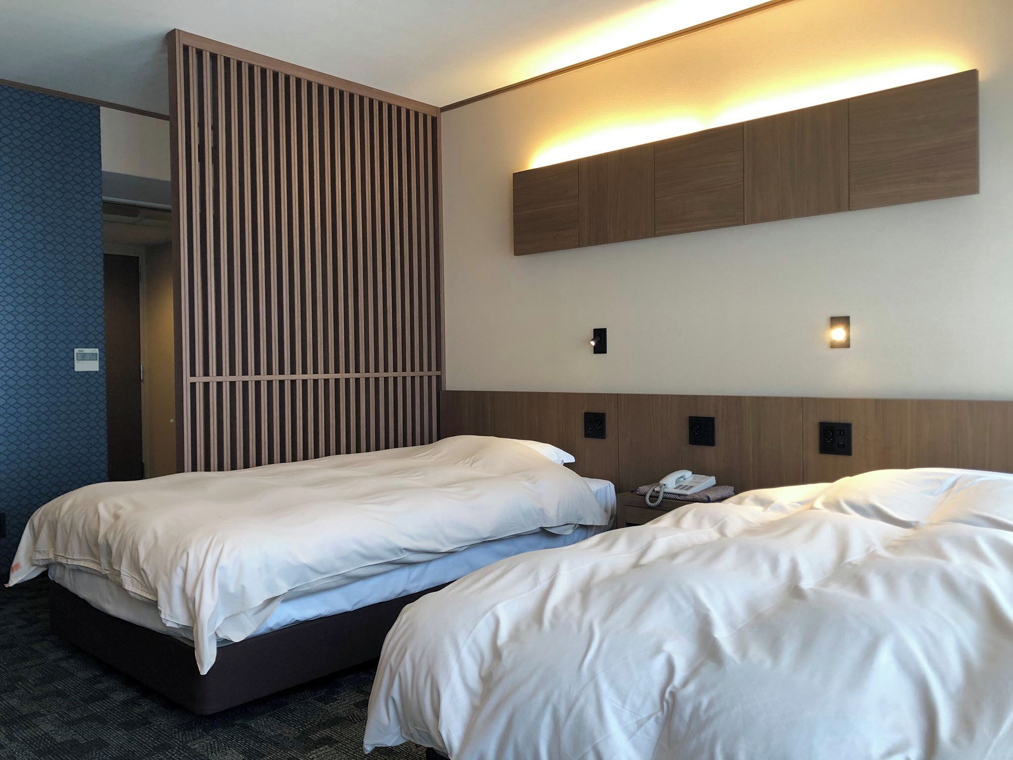 Ein Hotelzimmer mit zwei Betten, einer vertikalen Holzverkleidung, einem an der Wand montierten Telefon und einer Lampe.