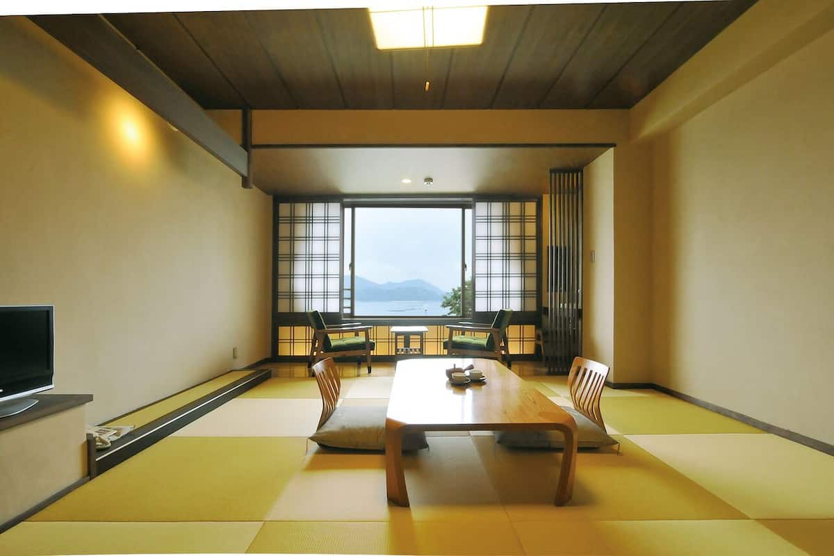 A room with tatami flooring, a wooden table, chairs, a television, and large windows with a view of the sea.