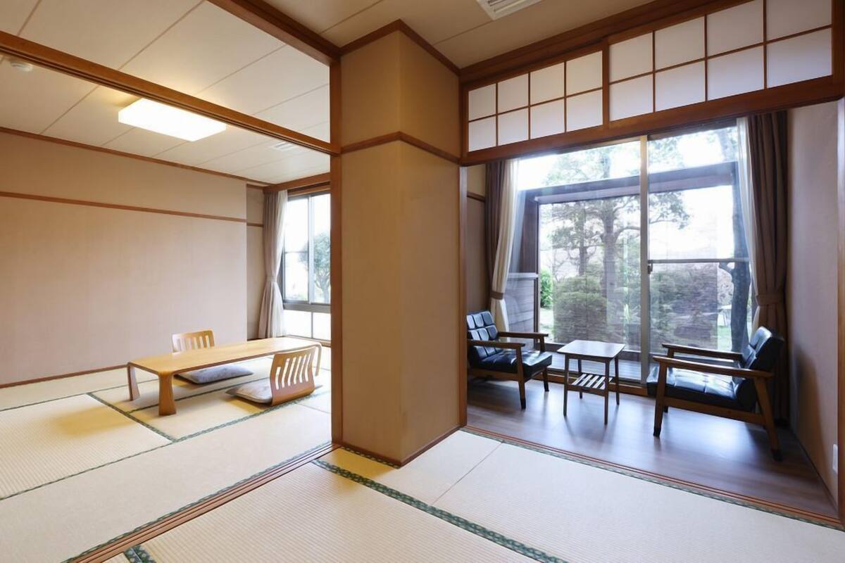 A traditional Japanese room with tatami flooring, a low table, chairs, and a large window with a view of trees.