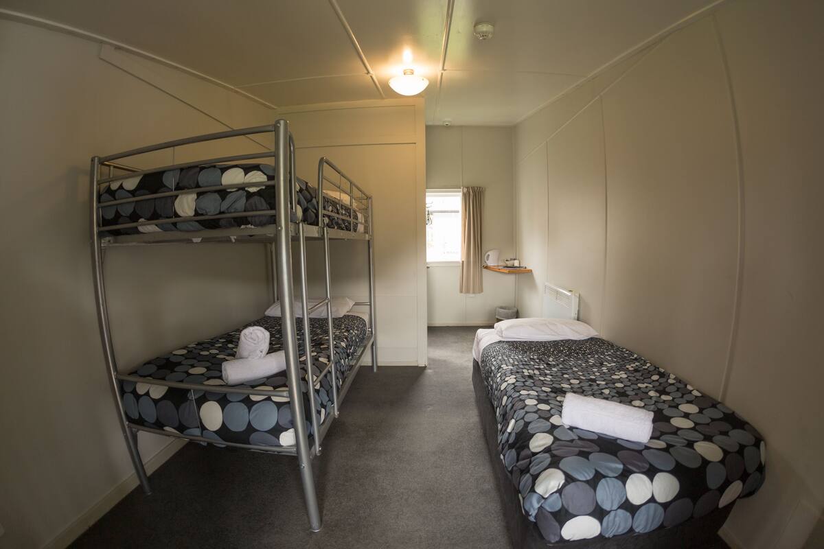 A dormitory room with bunk beds, a small kitchenette, and a window with curtains.