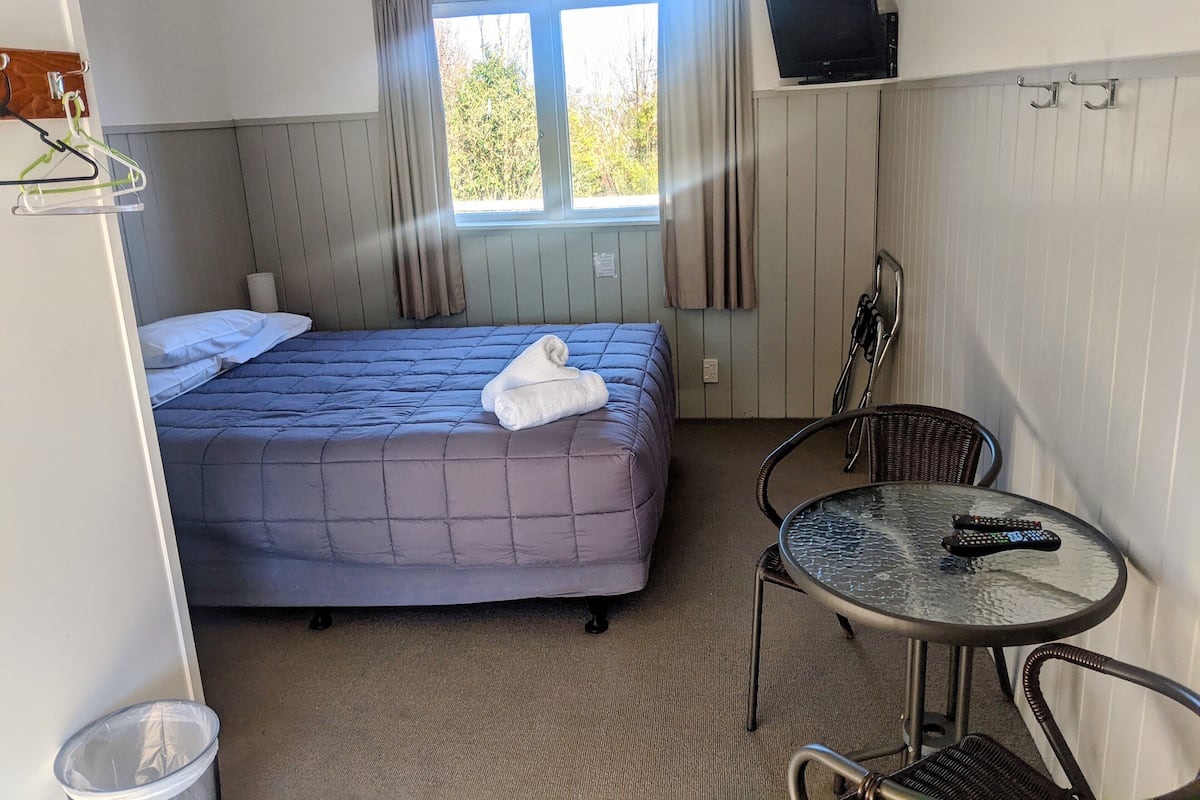 A hotel room with a bed, a TV on a stand, a table with chairs, and a window with curtains.